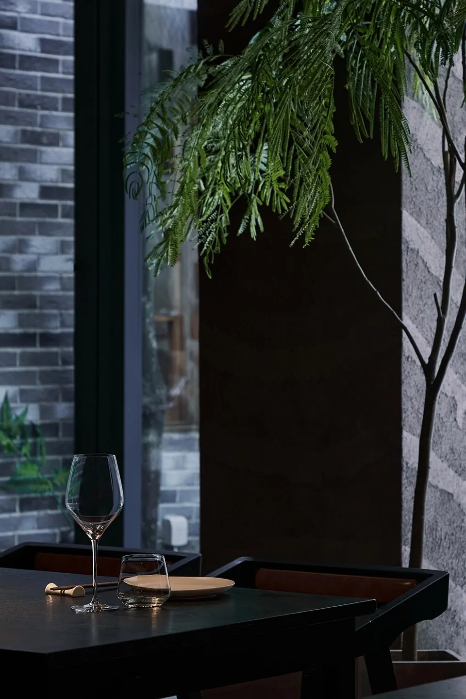 A sleek black dining table with a plant, glassware, and ceramic plates in an industrial-themed restaurant.