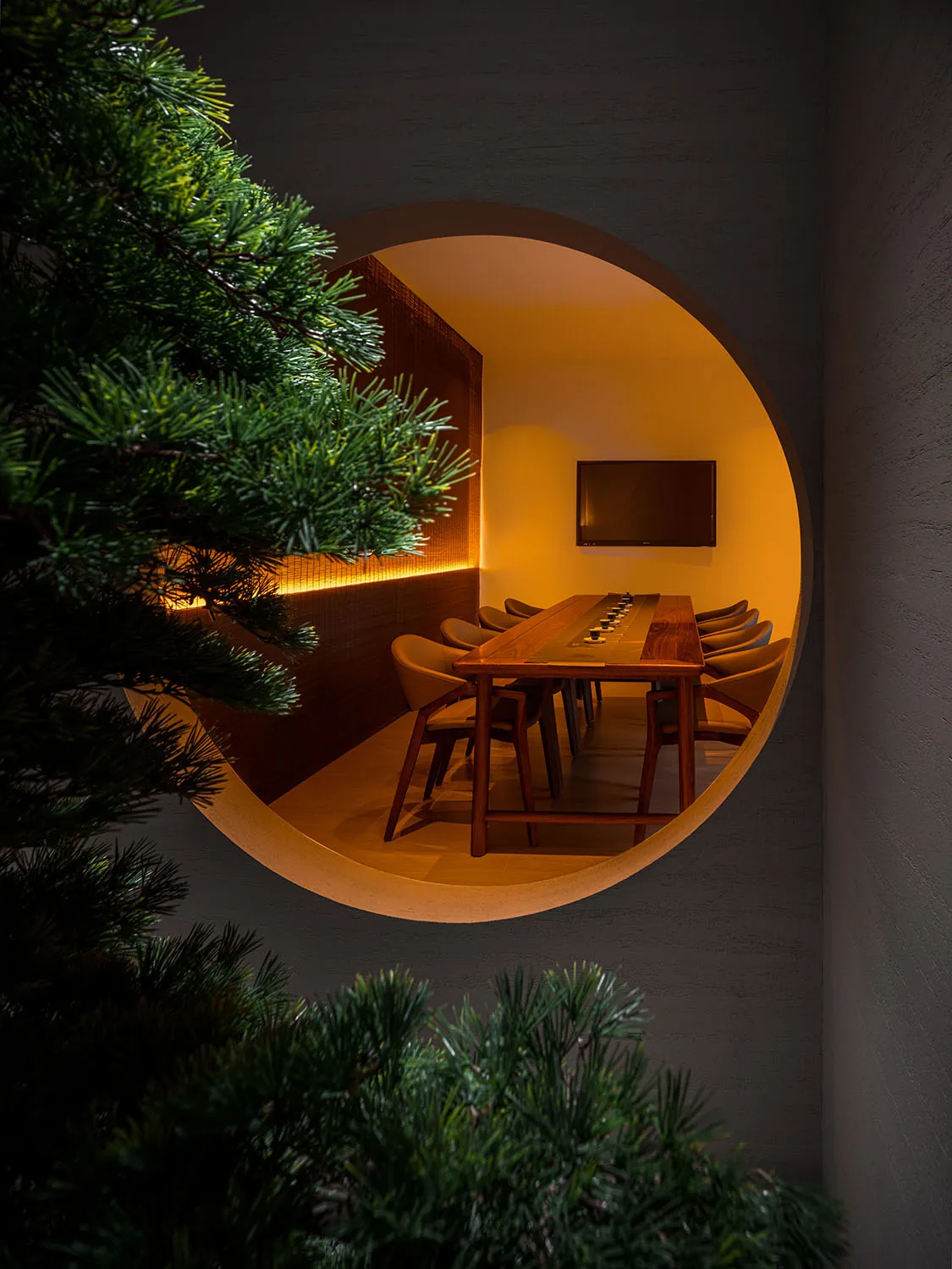A minimalist conference room featuring a long wooden table and chairs, with a round entryway and warm lighting.