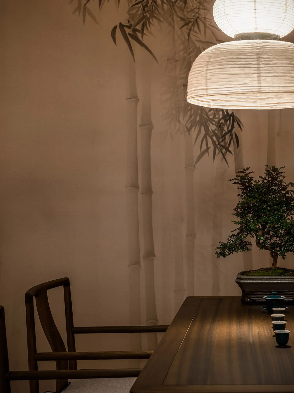 A minimalist table setting with a wooden table, a bonsai tree, tea cups, and a paper lamp.