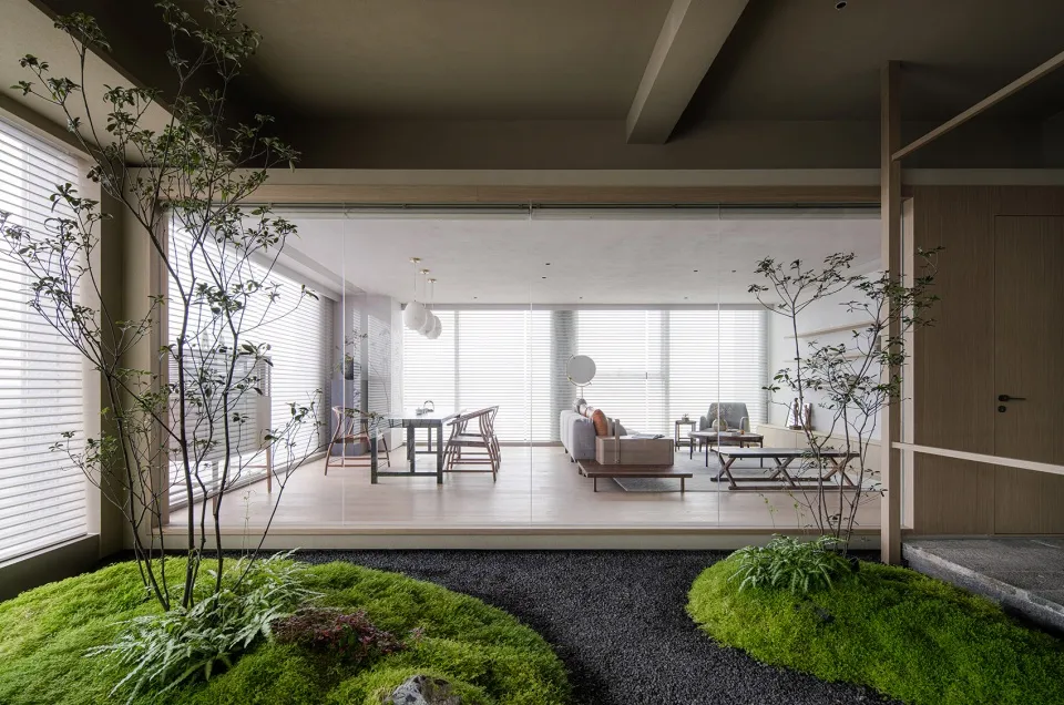 A modern minimalist living room features a glass coffee table on a wooden platform, surrounded by green plants and natural li