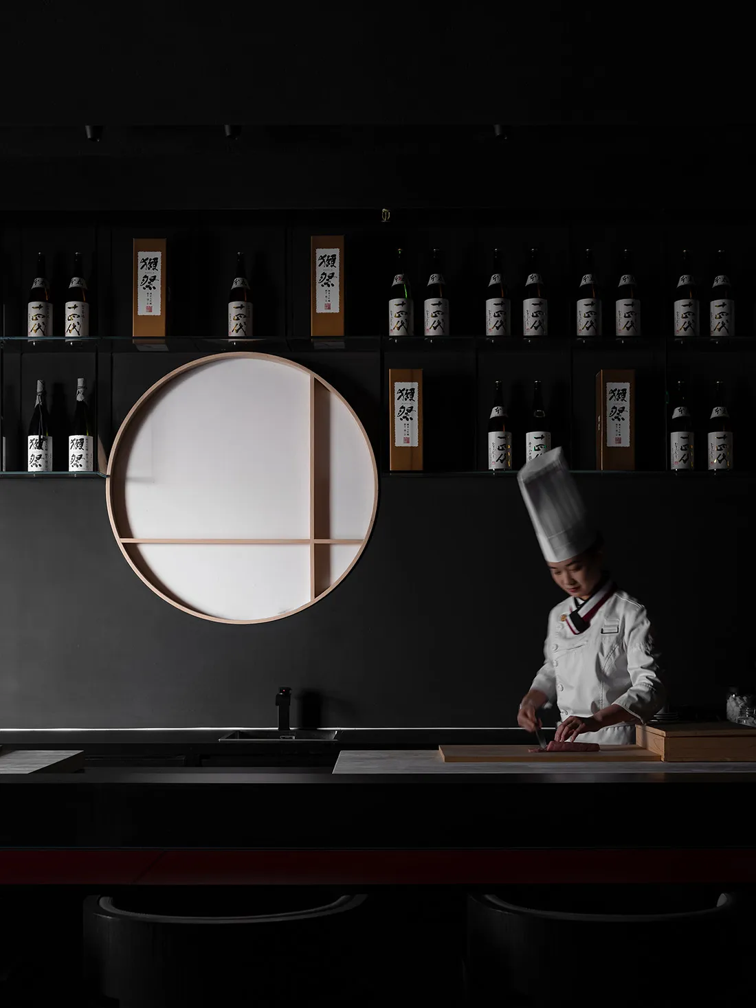 A minimalist restaurant interior with dark walls and a white table, featuring a chef preparing food.
