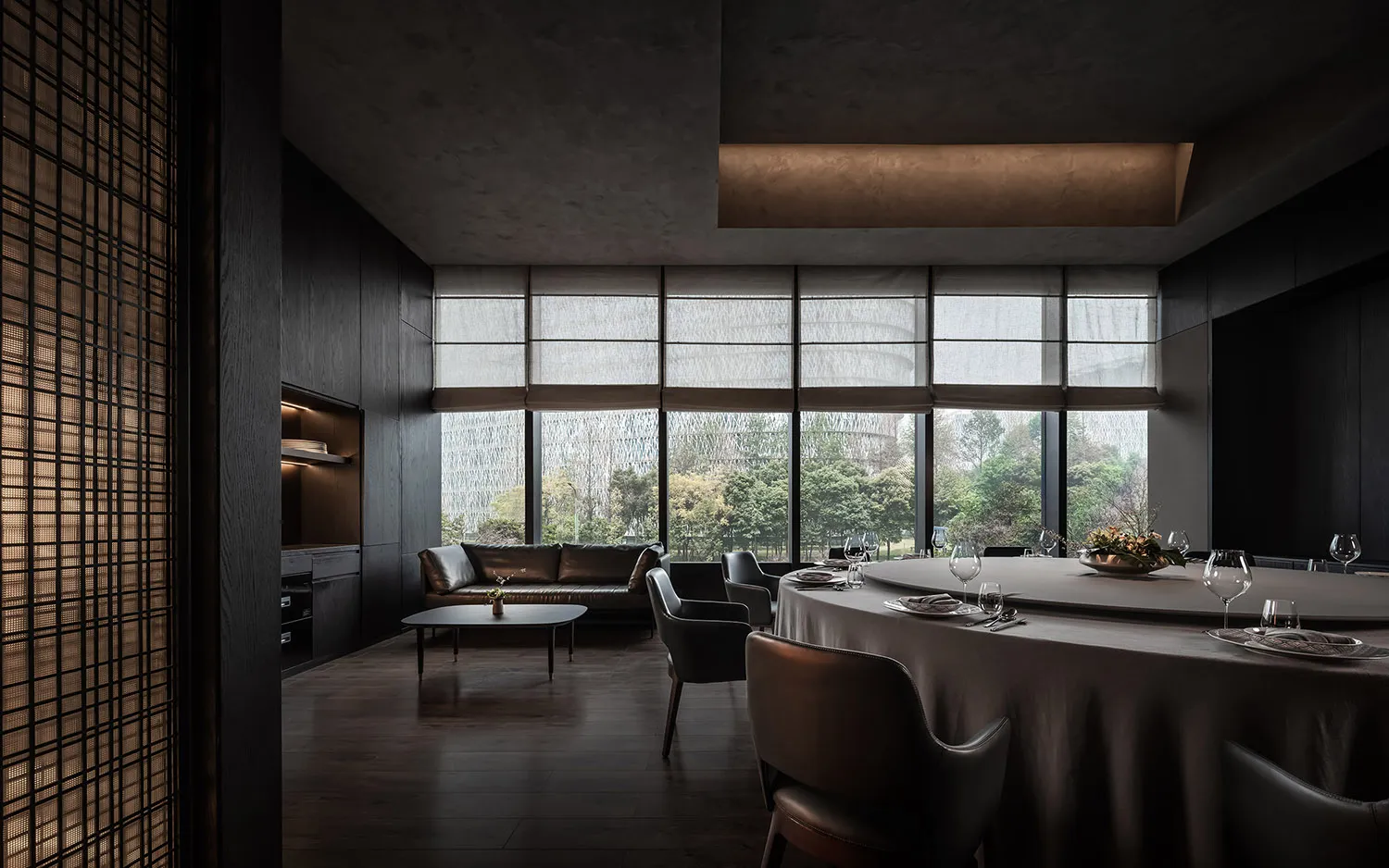 Modern restaurant dining room with round table, chairs, and large windows overlooking greenery.
