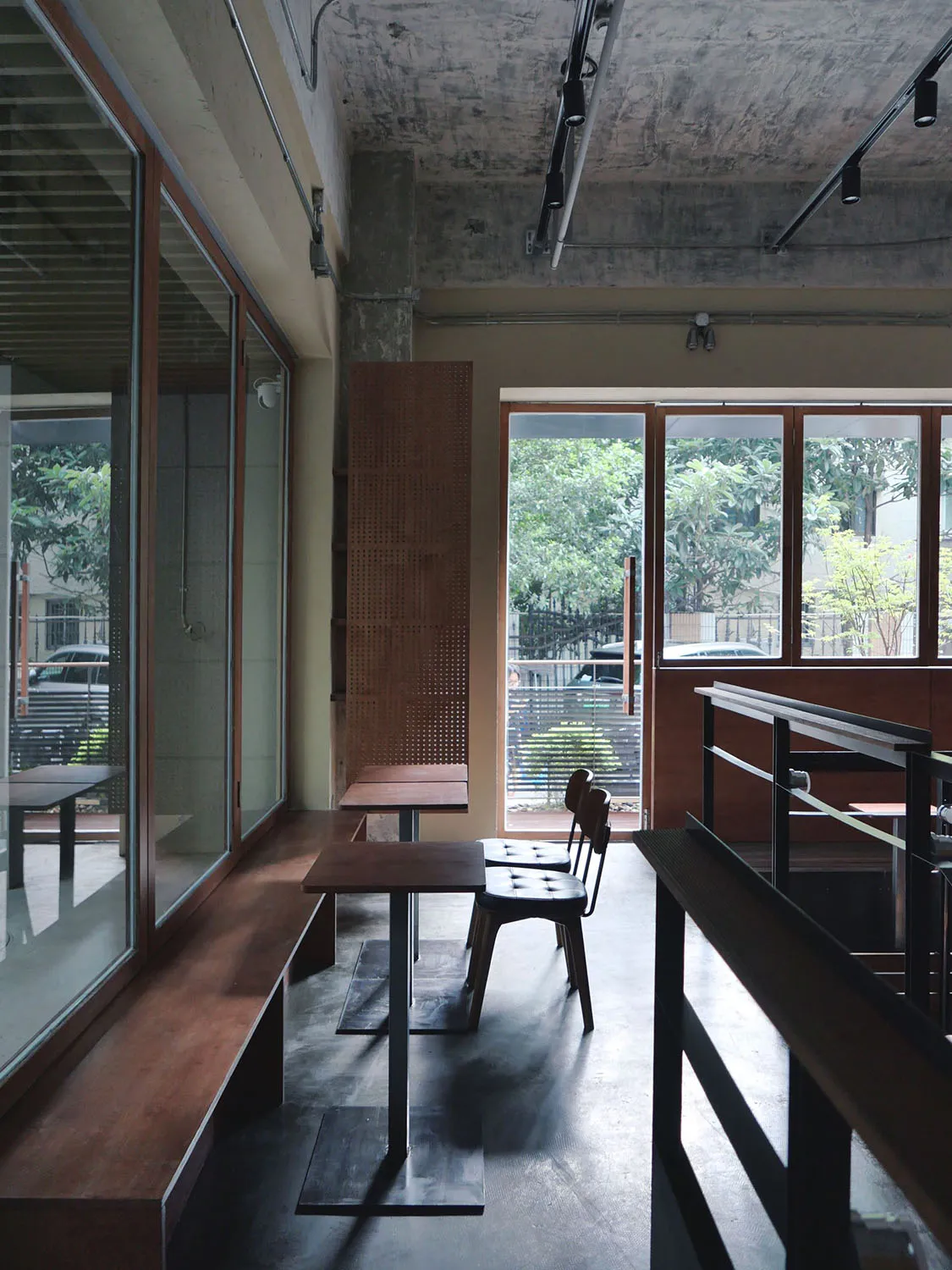 A modern cafe interior with wooden tables and chairs, concrete flooring, and large windows showing greenery outside.