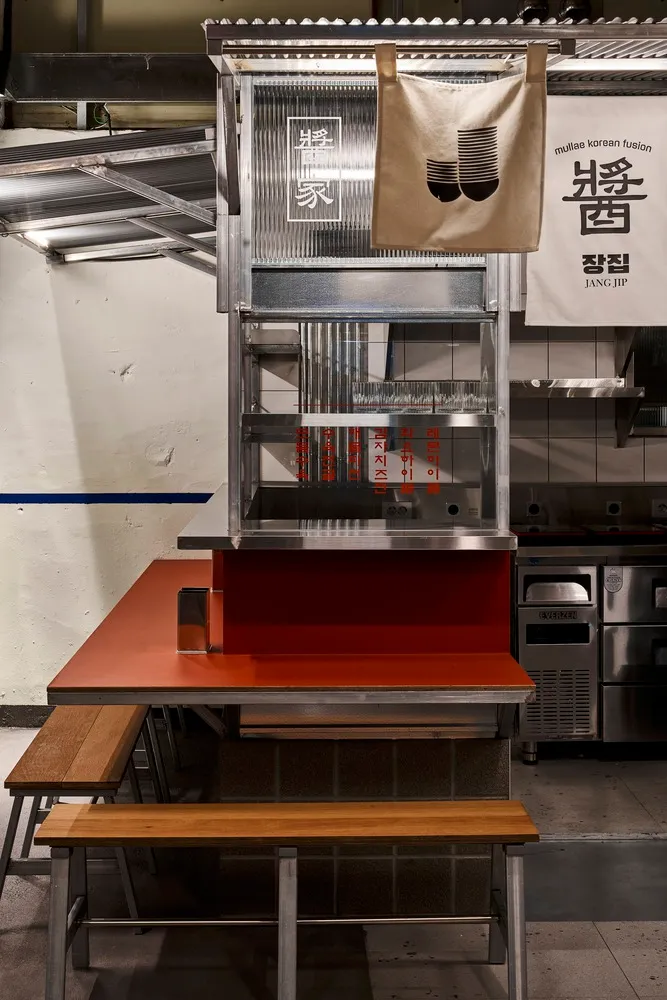 A minimalist restaurant interior with a metal food counter, wooden benches, and Korean signage.