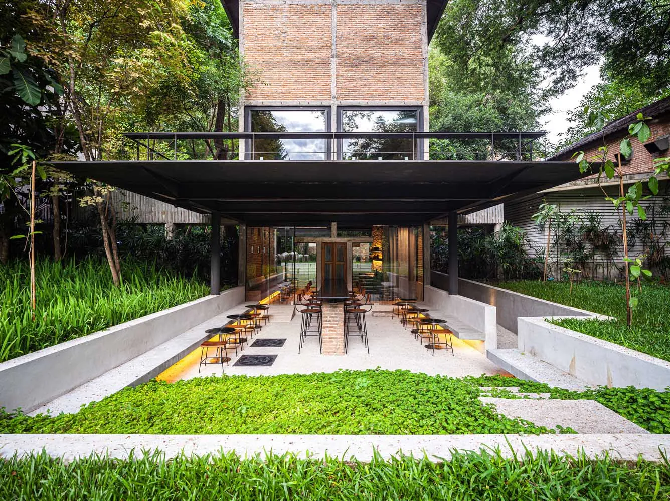A modern restaurant exterior features a brick façade with a concrete patio surrounded by green plants.