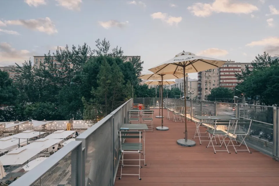 Modern rooftop terrace featuring wood flooring with tables and chairs, surrounded by trees and cityscape.