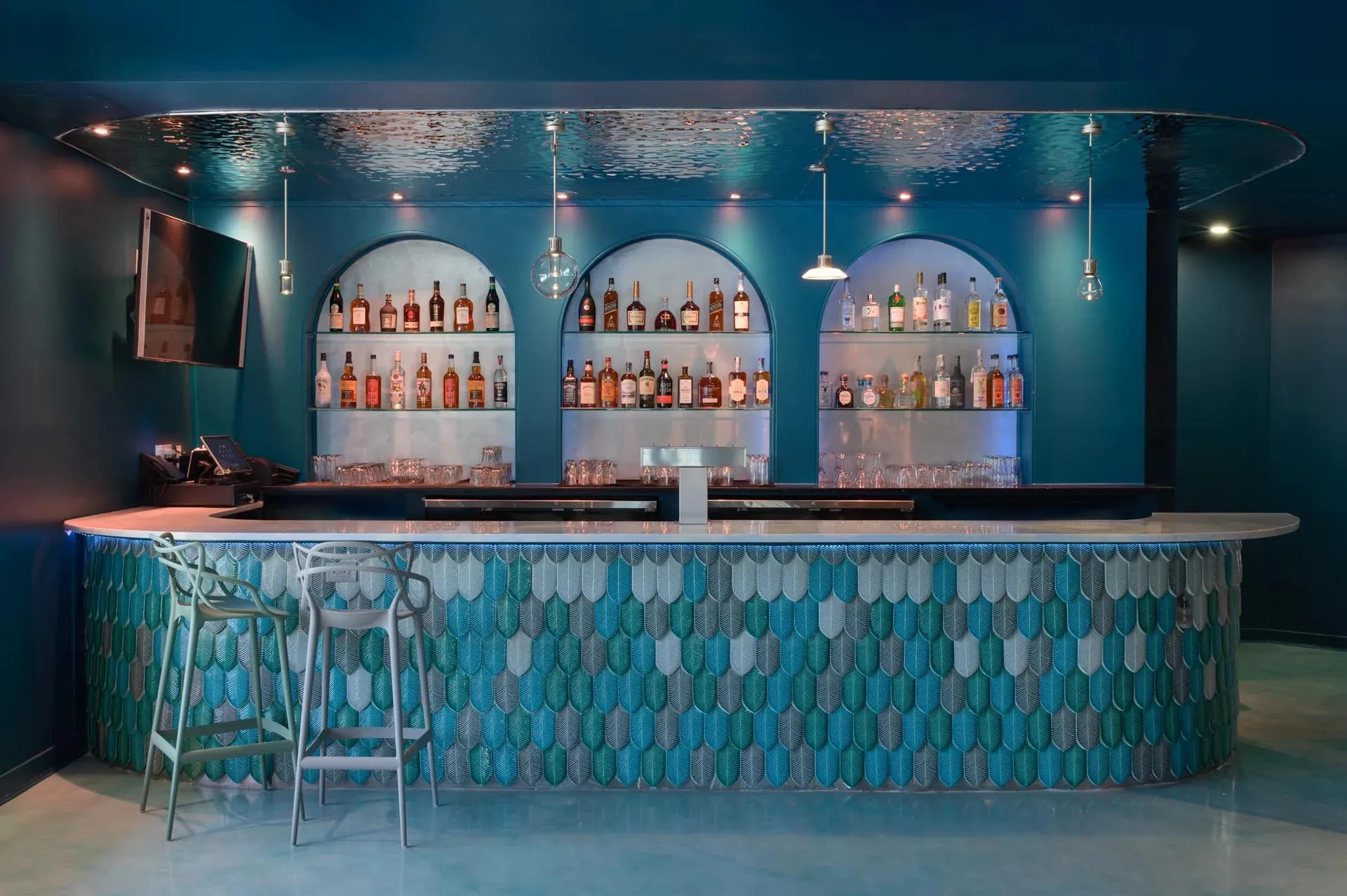 A modern bar with blue ceramic tile walls, a curved countertop, and various bottles of alcohol on shelves.