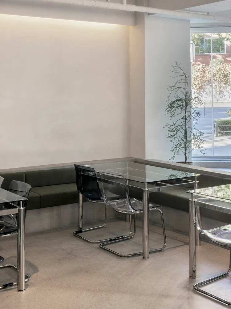 A modern industrial restaurant interior featuring glass tables, concrete bench seating, and a canvas wall.