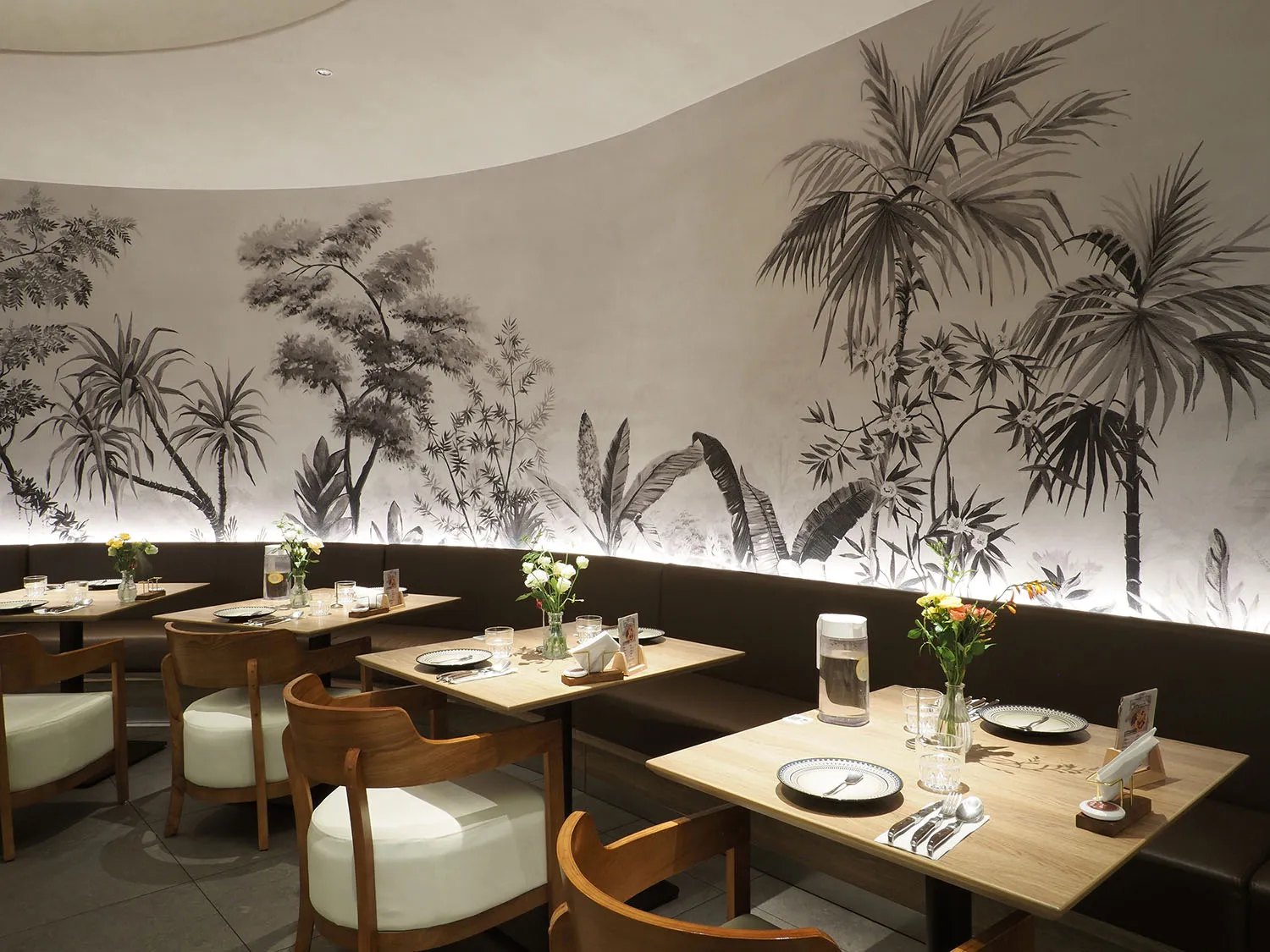 Wooden tables and chairs in a cafe interior with a mural of tropical plants on the wall.