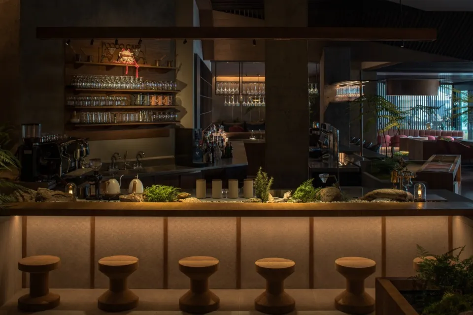 A modern bar interior featuring wooden stools and concrete columns, with a well-stocked bar counter and greenery on the count