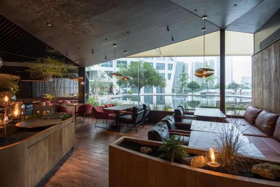 A modern restaurant with wooden furniture, plants, and a view of the city through large windows.