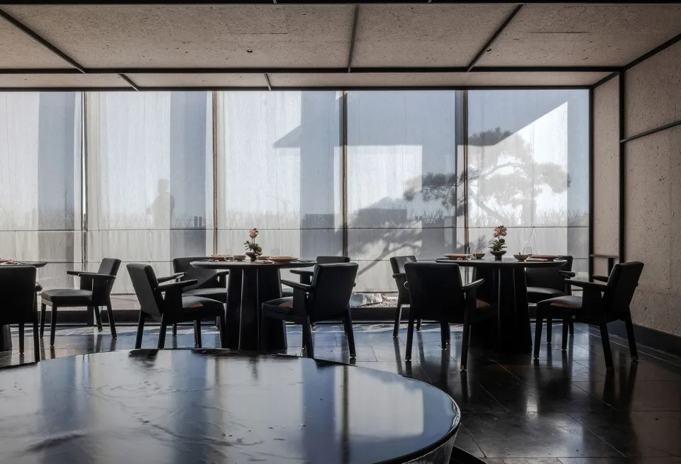 現代餐廳室內空間,配有黑色餐桌、餐椅與地板,大型窗戶搭配薄紗窗簾,可眺望窗外樹木