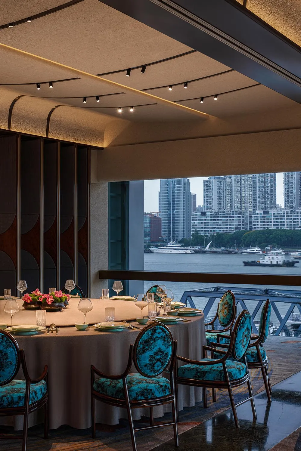 A modern restaurant interior with teal upholstered chairs, round tables, and a view of the cityscape.