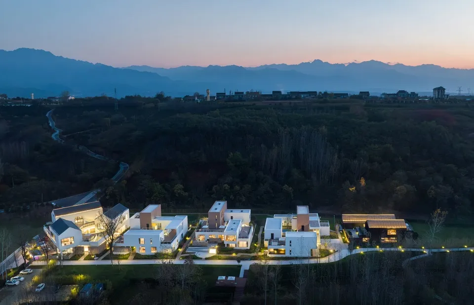 Aerial view of a modern residential housing community during twilight with illuminated buildings and mountainous background.