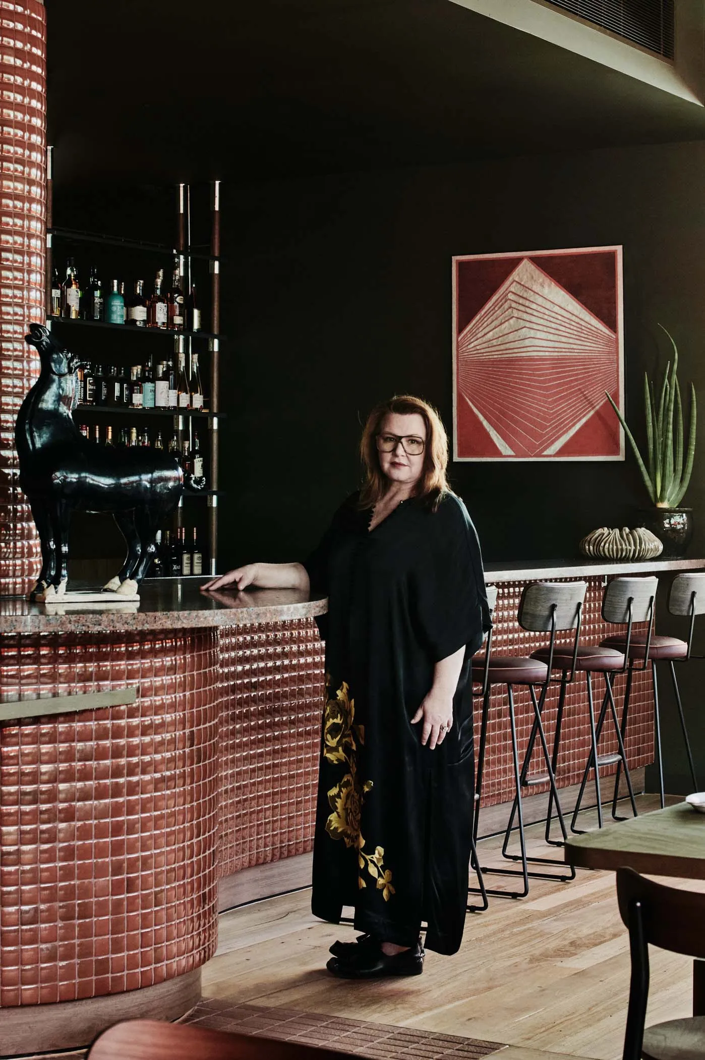 A woman standing at a modern restaurant with a red tile bar counter, black stools, and a geometric artwork on the wall.