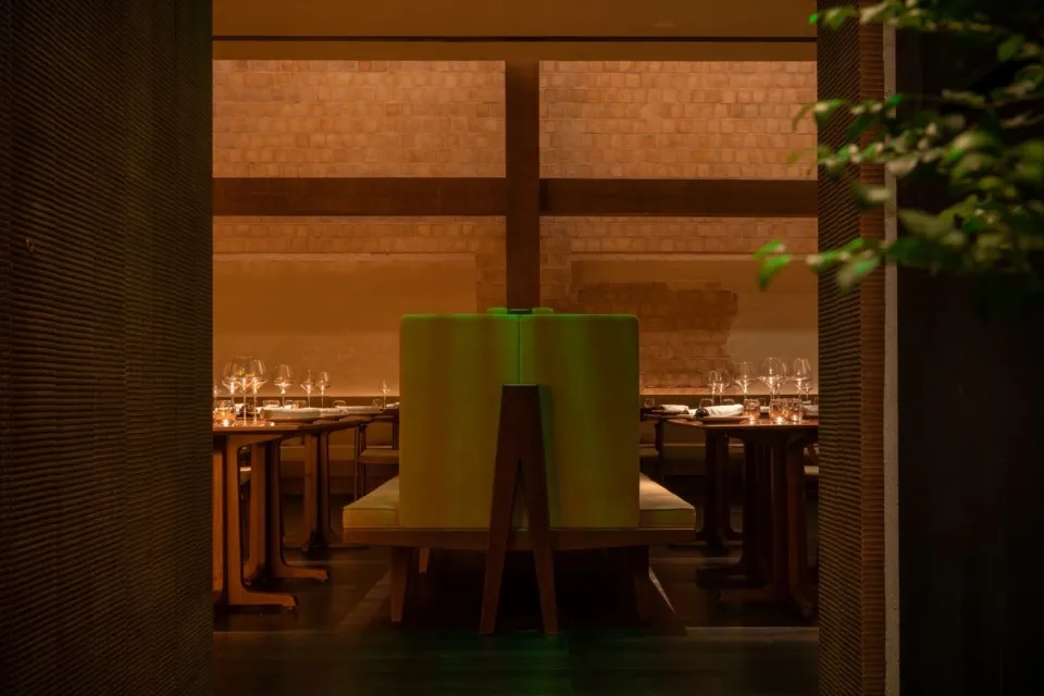 Modern restaurant dining room with green chairs and wooden tables, illuminated by soft lighting and surrounded by greenery.