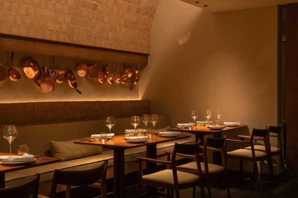 A cozy restaurant interior with dark wood tables and chairs, warm lighting, and hanging copper pots.