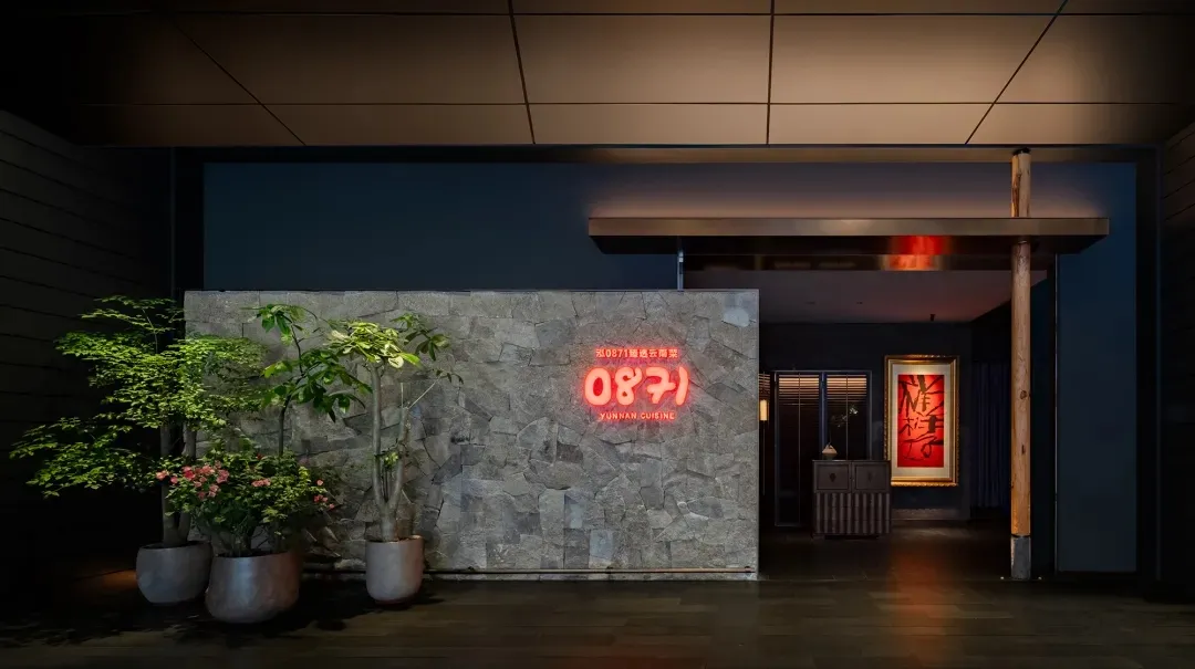 A modern restaurant interior with concrete walls, plants, and a neon sign.