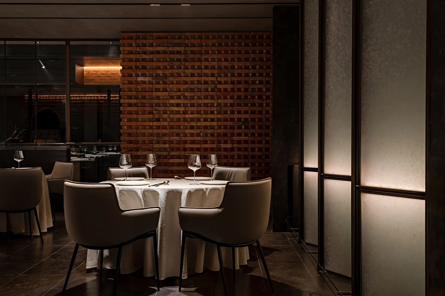 A dimly lit dining room with a brick wall, round table set for four, and sleek, modern chairs.