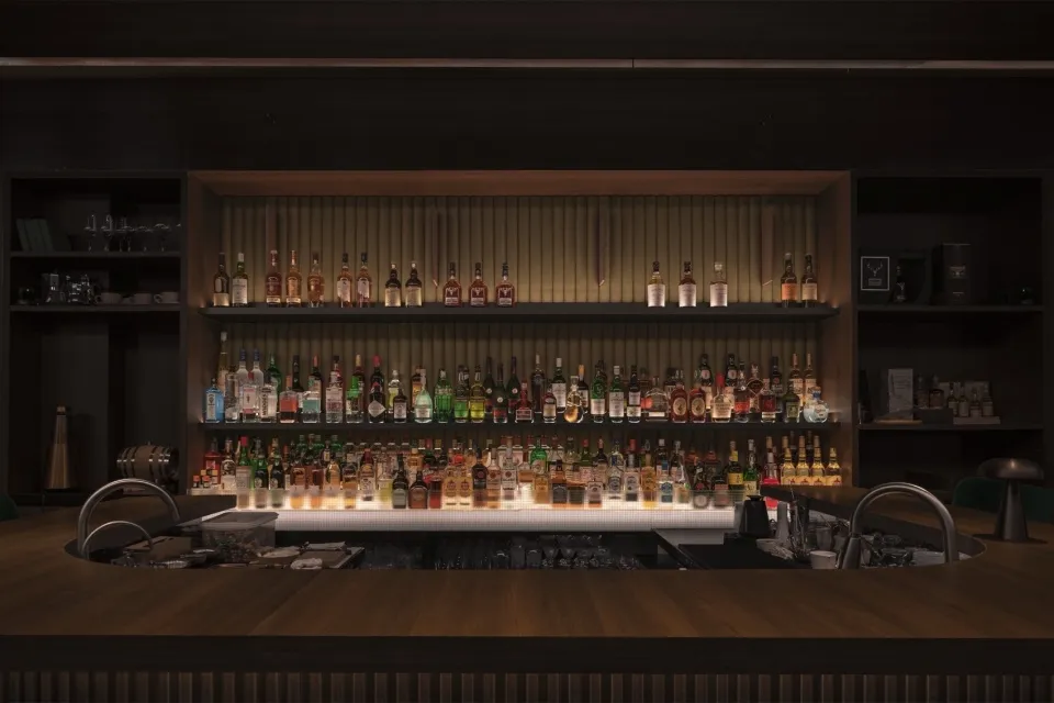 A modern bar counter made of wood, featuring a variety of bottles on shelves.