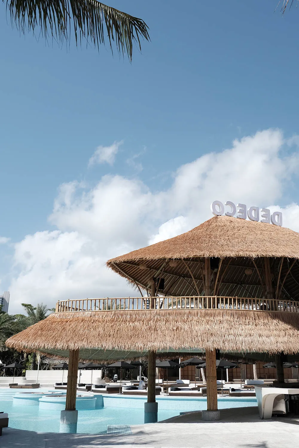 A modern, thatched-roof poolside bar under a clear blue sky with white clouds.