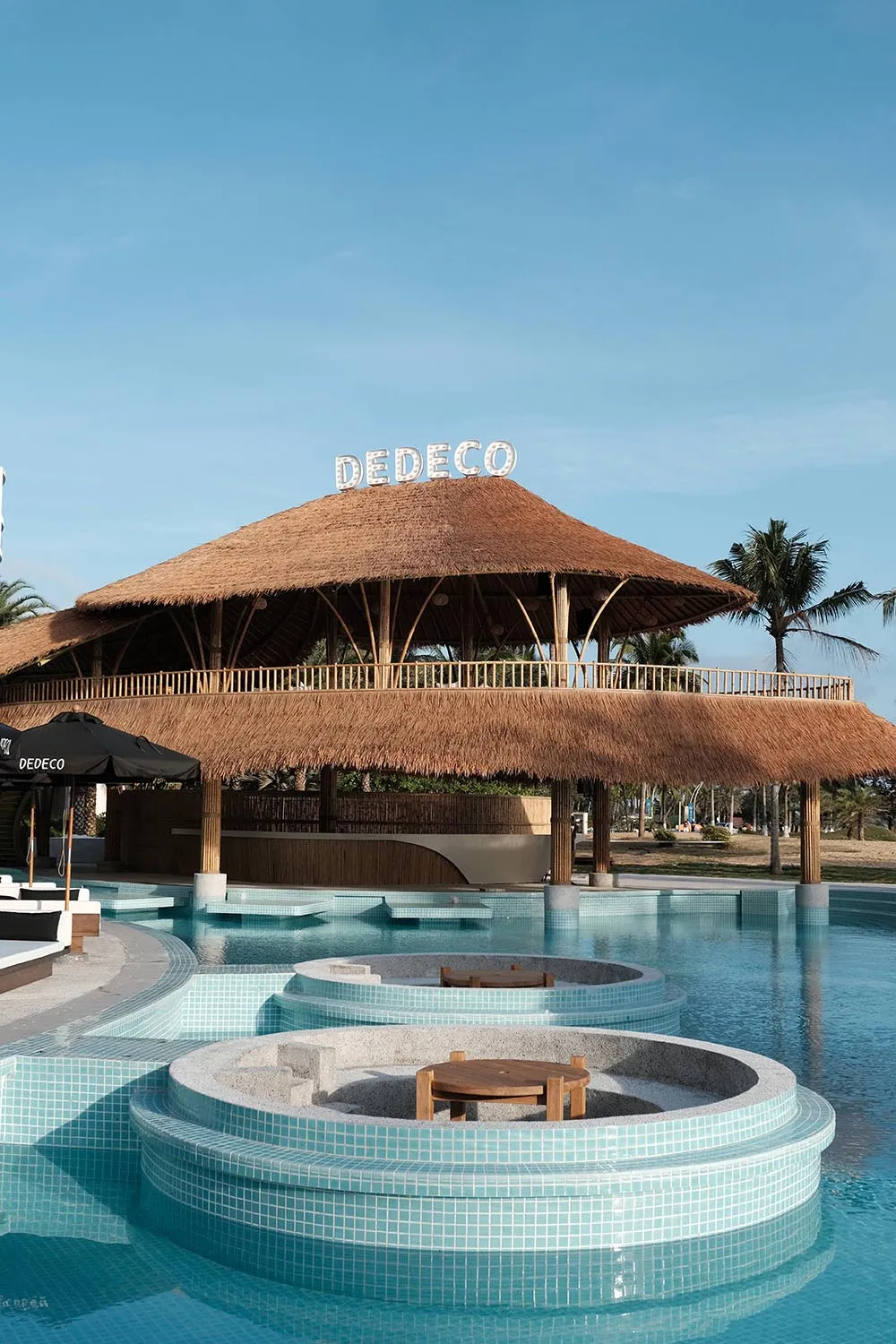 A poolside lounge area featuring a thatched-roof pavilion with wooden columns and seating.