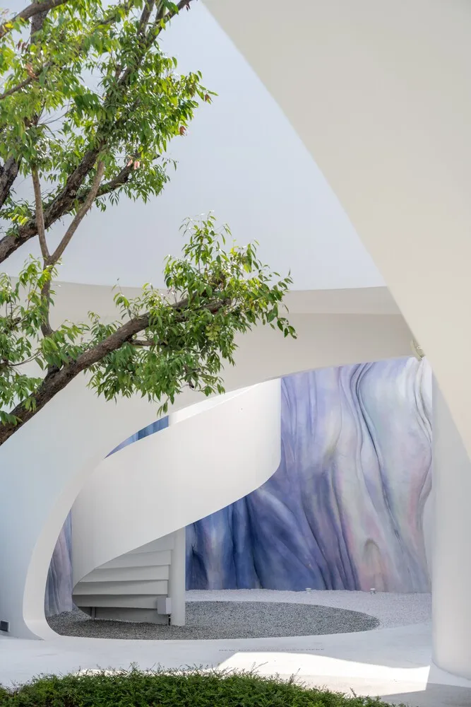 An outdoor modern architectural space featuring a white curved staircase and wall with a mix of blue and purple hues.