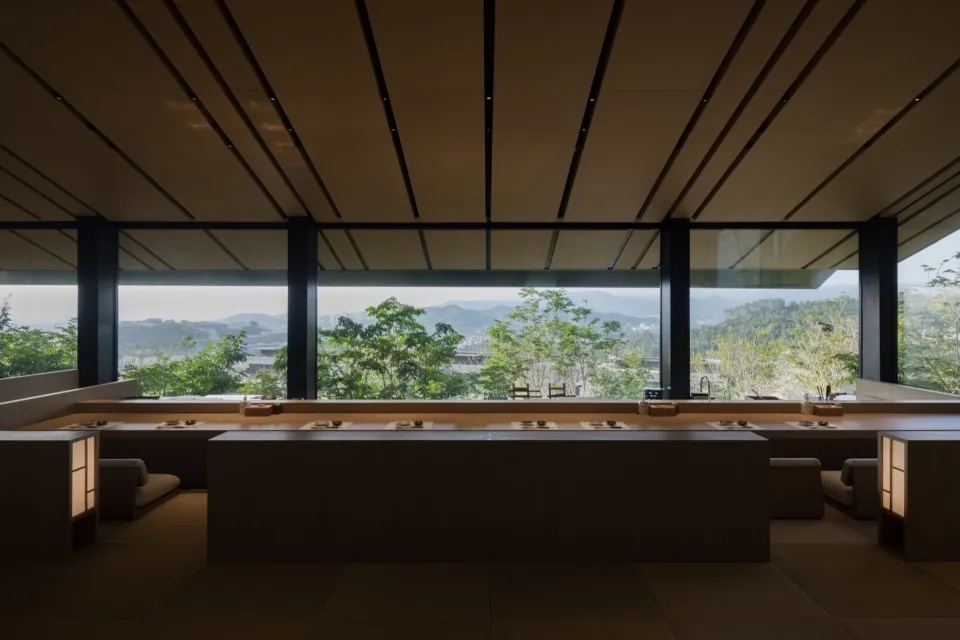 Modern restaurant interior with wooden tables, concrete floor, and floor-to-ceiling windows overlooking natural scenery.