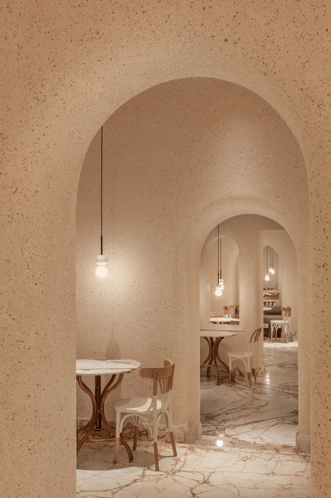 A modern restaurant interior with concrete walls and floor, featuring minimalist furniture and geometric light fixtures.