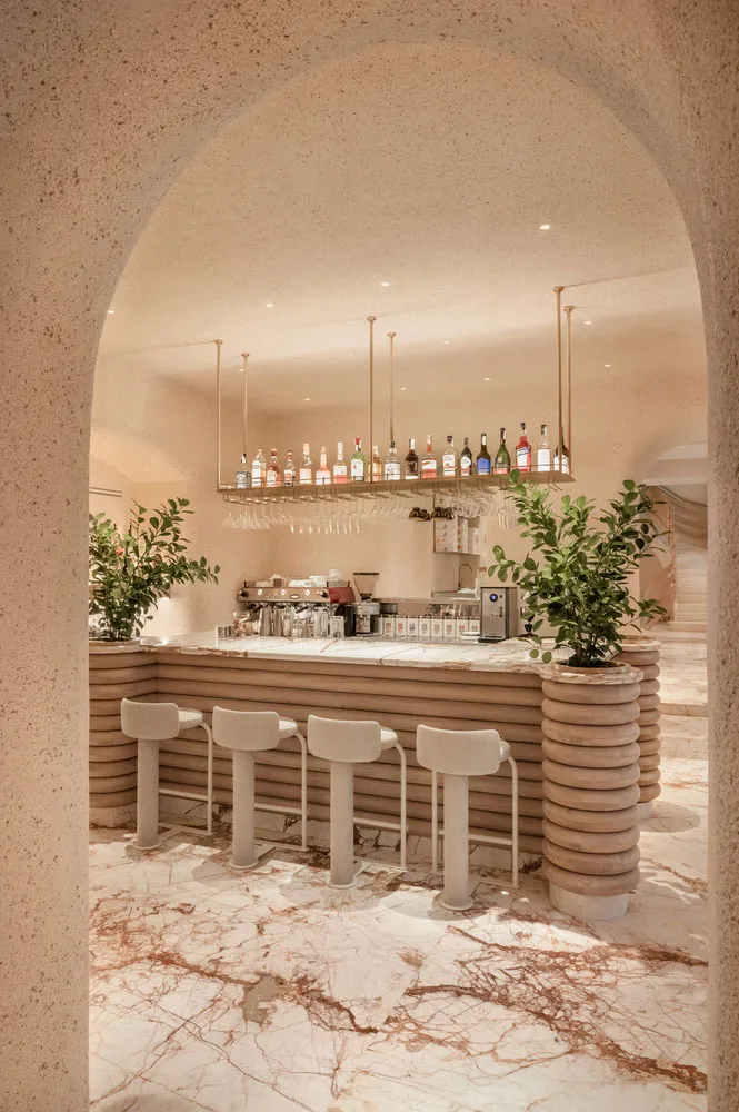 A modern bar interior with marble and concrete materials, featuring a counter, stools, and a shelf with various bottles and g
