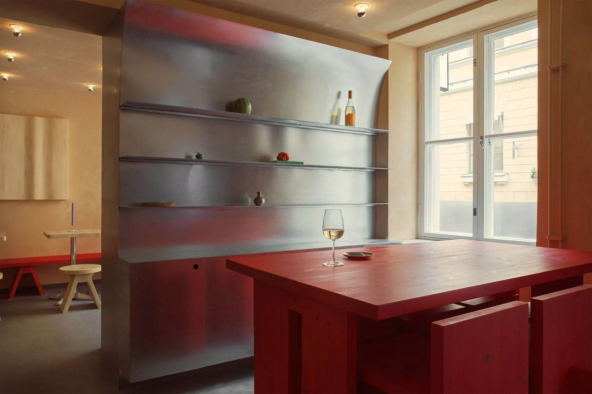 A minimalist dining room with a red wooden table and chairs, featuring a metallic shelving unit and a window with a view of a
