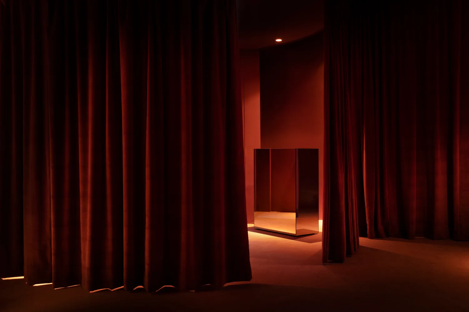 Dark red velvet theater stage curtains in front of a dimly lit, minimalist interior with a mirrored box in the center.