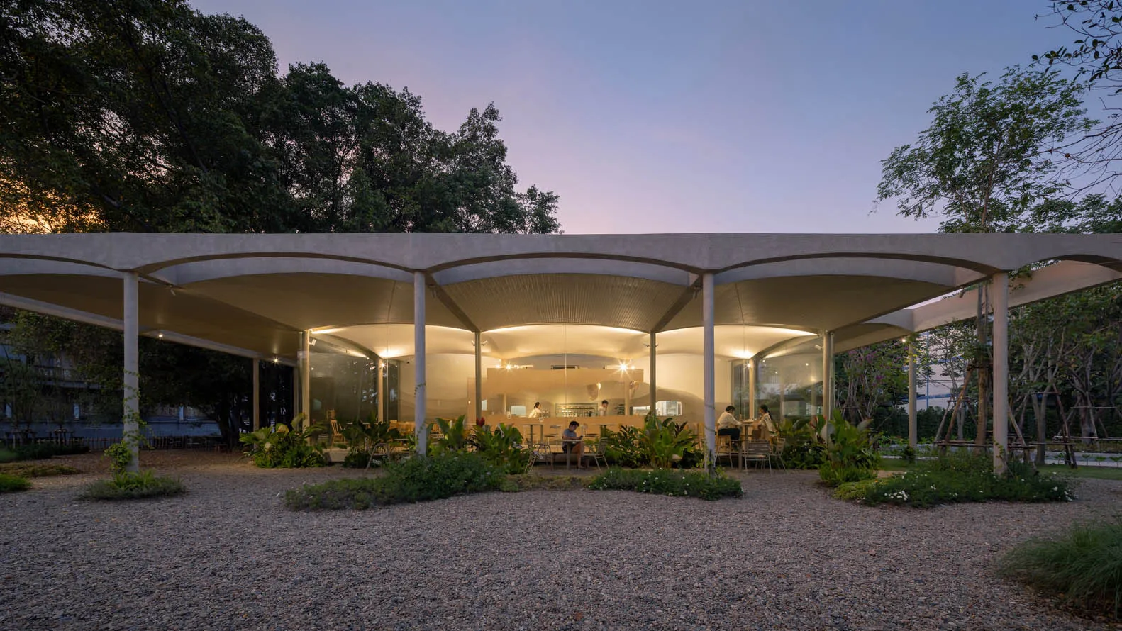 An illuminated restaurant with a modern design, featuring concrete structures and greenery.