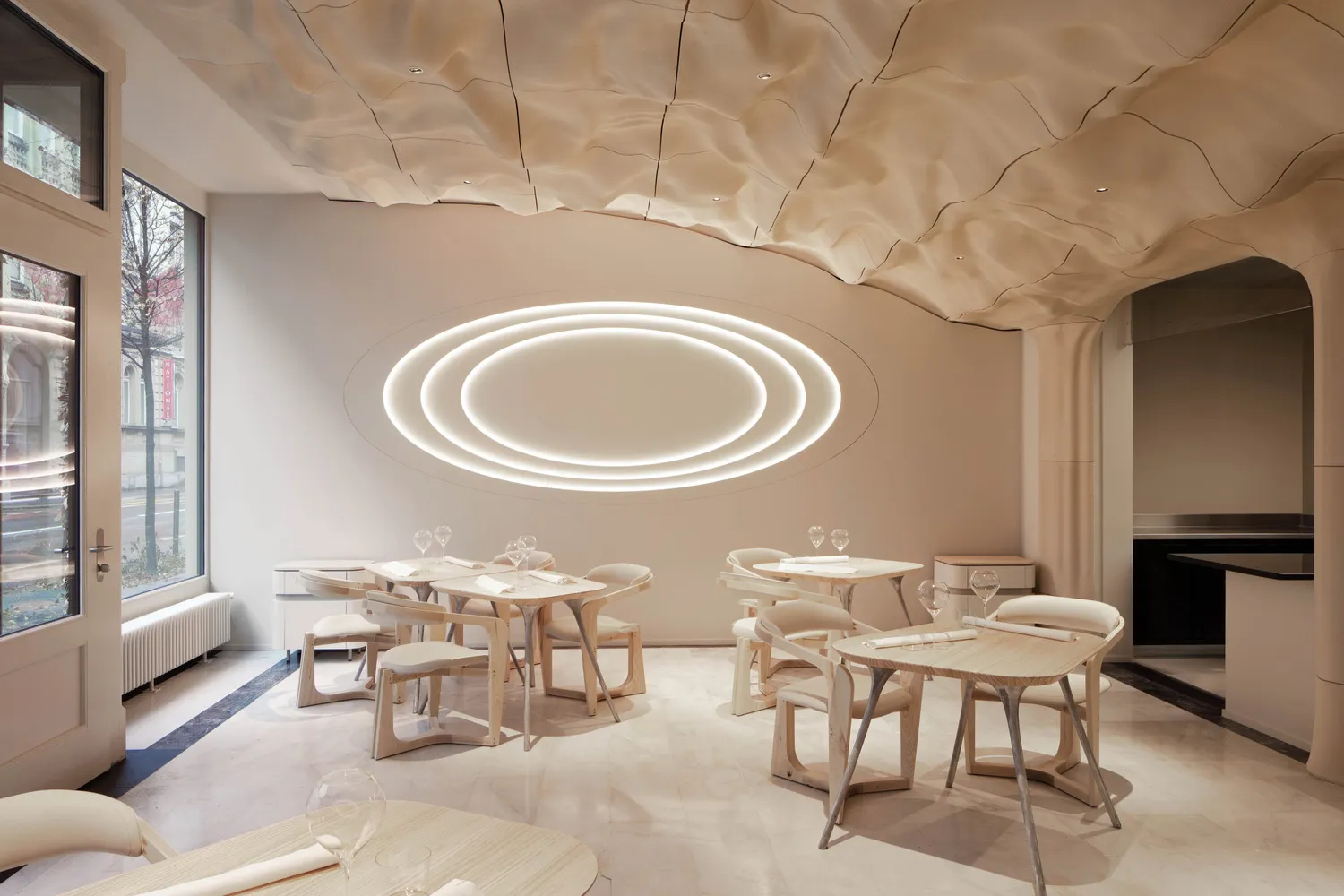 A modern restaurant dining room with white walls, circular lighting fixtures, and wooden tables and chairs.