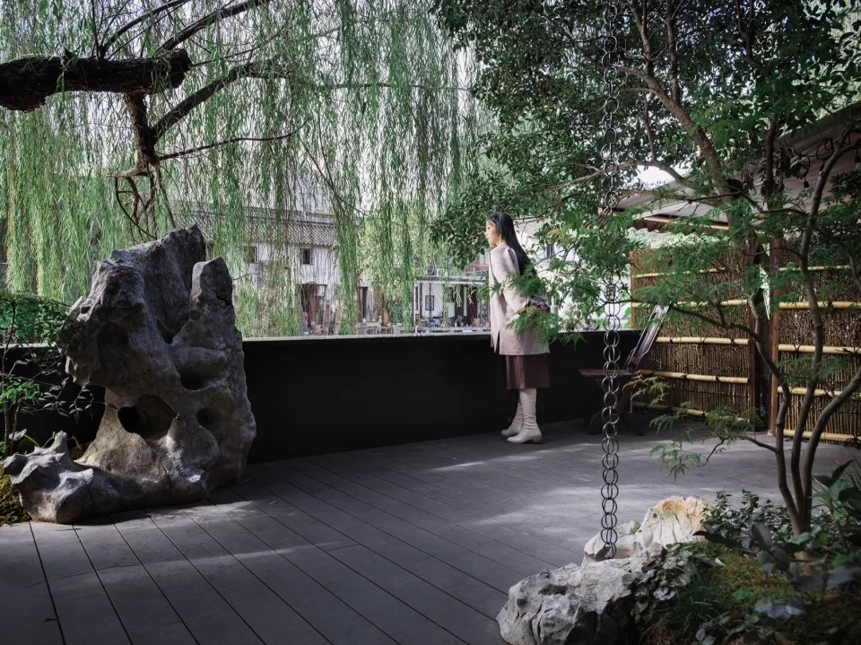 A serene garden with stone formations and greenery, featuring a woman standing near a wooden fence and a swing.