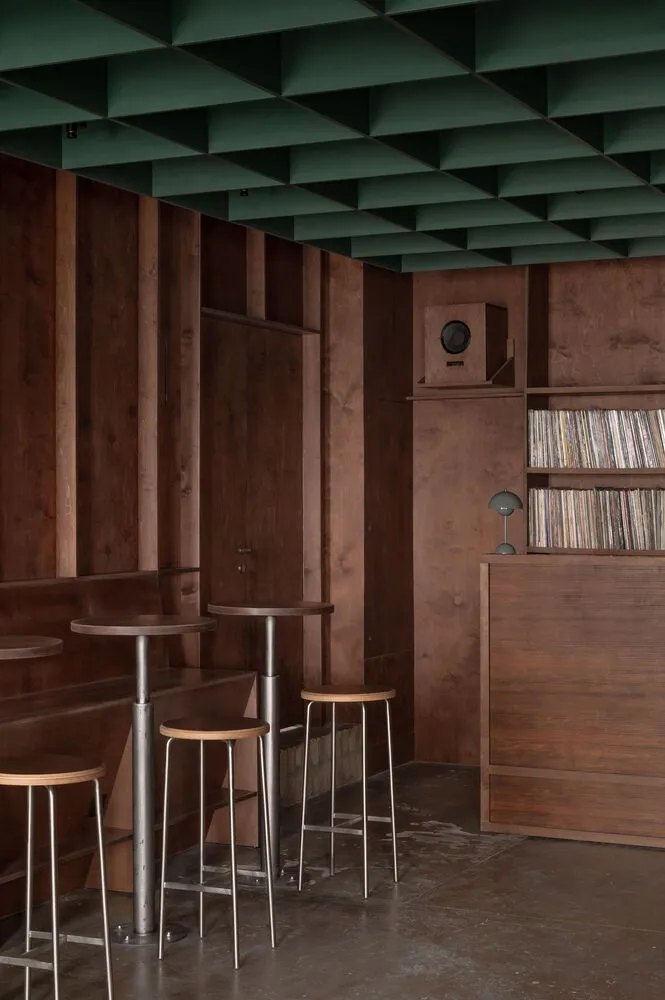 Industrial-style coffee shop featuring a wooden interior with high stools, bar tables, and vinyl record shelves.