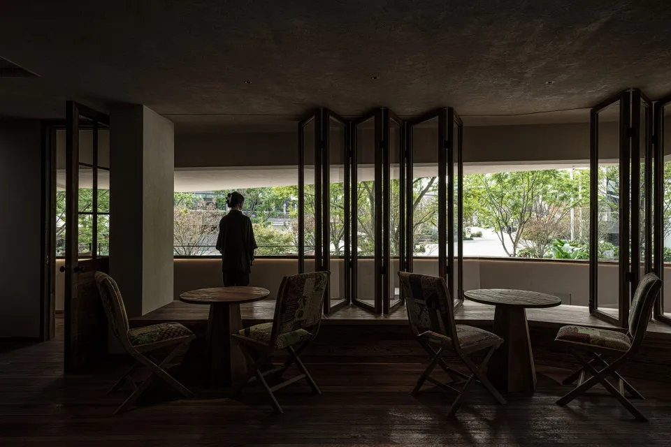 A coffee shop interior with wooden tables, concrete floors, and large glass windows overlooking greenery.