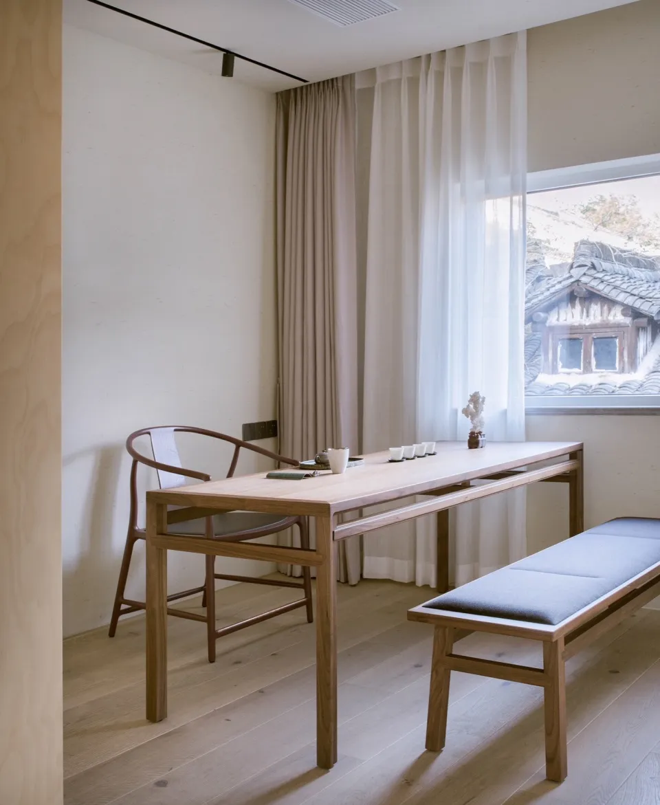 A minimalist interior featuring a wooden table and bench near a window with curtains and a wooden chair.