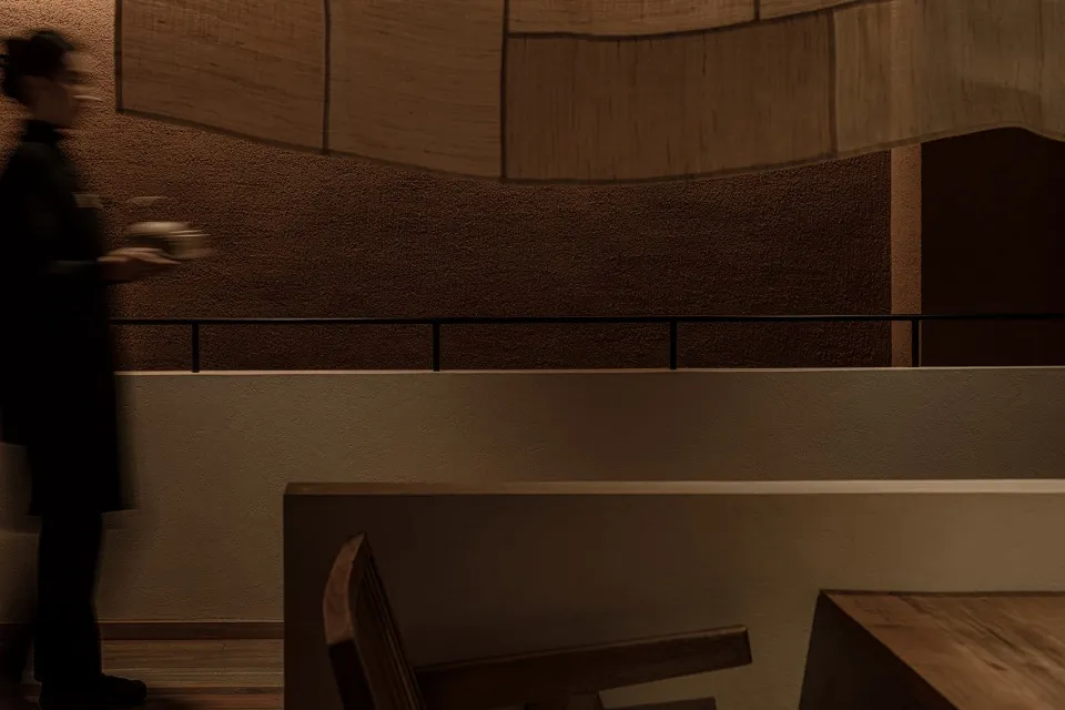 A person wearing a uniform carries a dish in a restaurant setting, featuring modern furniture and wood elements.