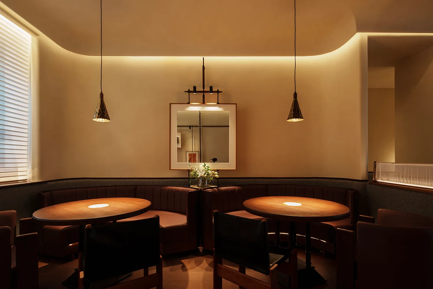 A modern restaurant dining room features round wooden tables with pendant lights, upholstered benches with leather accents, a