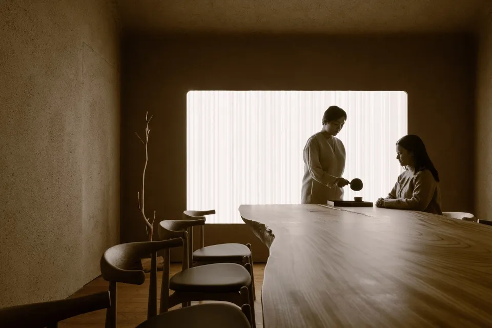 A minimalist tea ceremony room with a wooden table and a woman serving tea