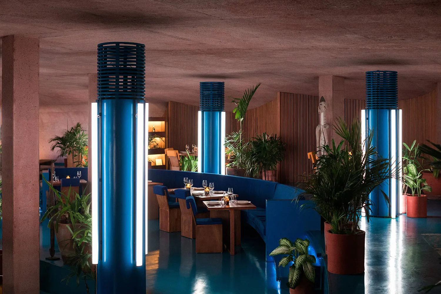 Interior view of a modern restaurant featuring blue light columns, concrete walls, and indoor plants.