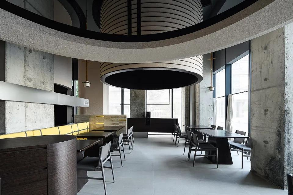 Modern restaurant interior with concrete walls, wooden furniture, and large windows.