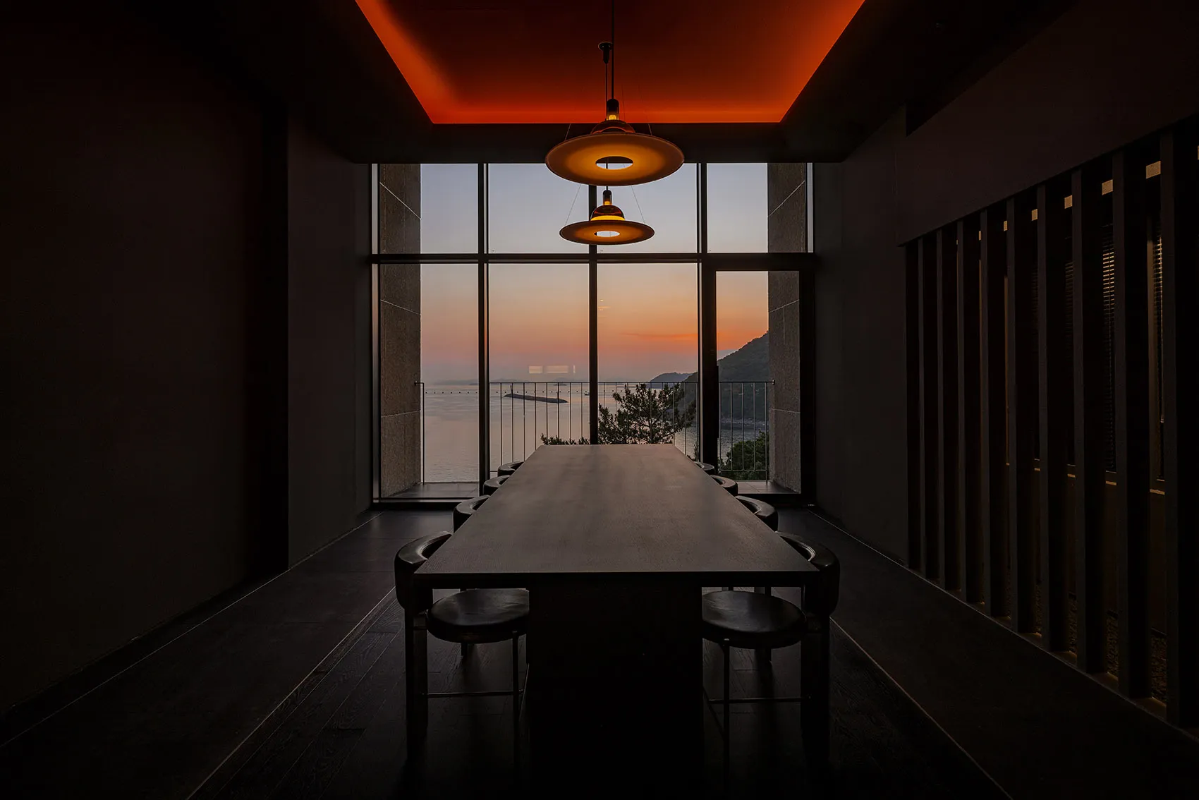 A modern dining room featuring a long wooden table and chairs, set against a large glass window overlooking a sunset over the