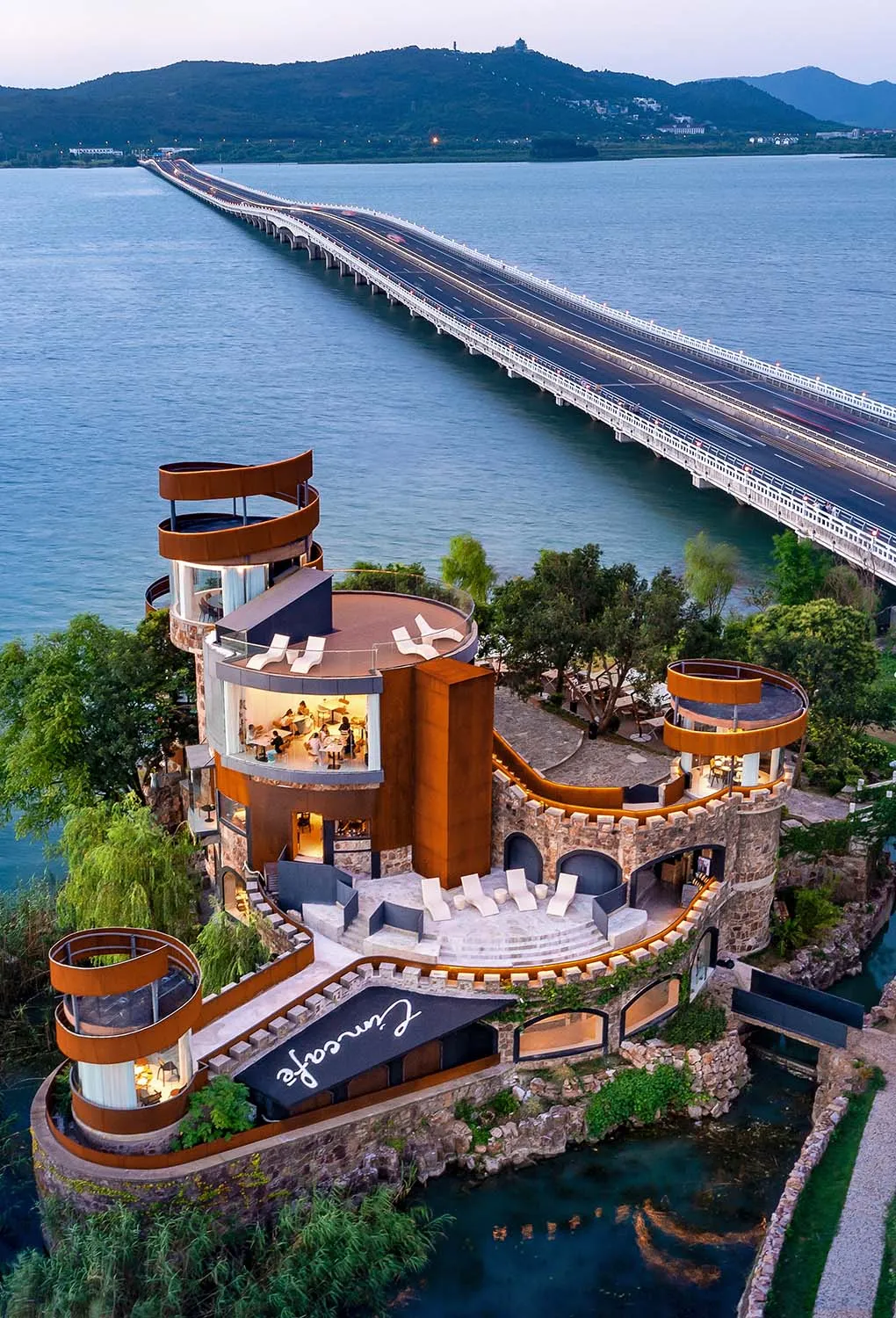 一座城堡式建築坐落於水面上，一條長橋延伸至遠方，背景為山巒。