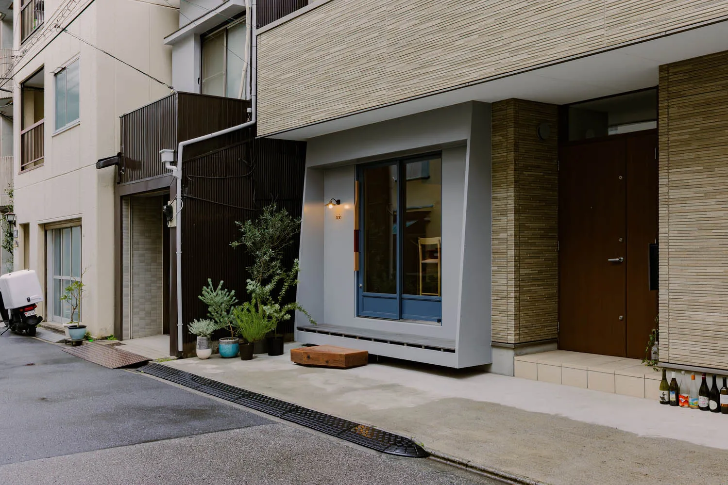 A modern storefront with a concrete exterior, featuring large windows, a wooden bench, and potted plants.