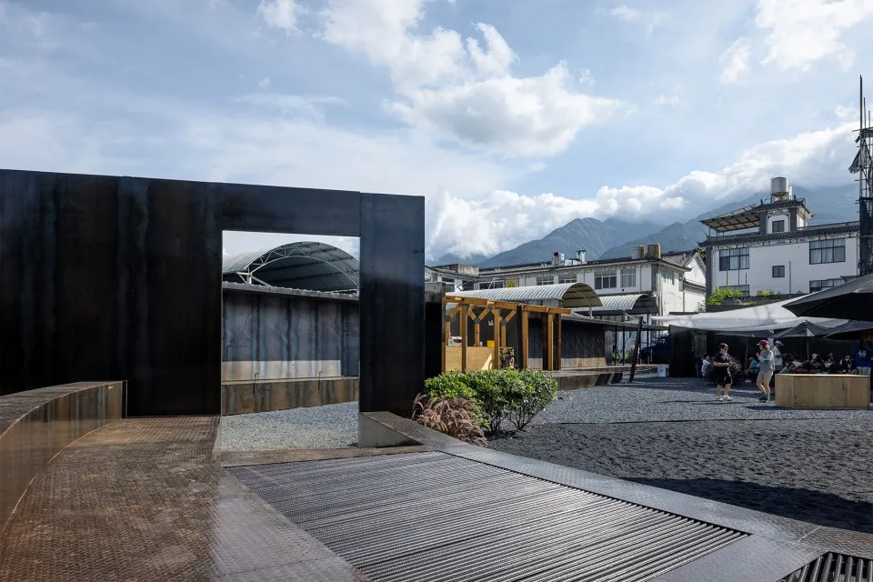 A minimalist urban architecture scene featuring a black CorTen steel block, concrete walkways, and wooden elements.