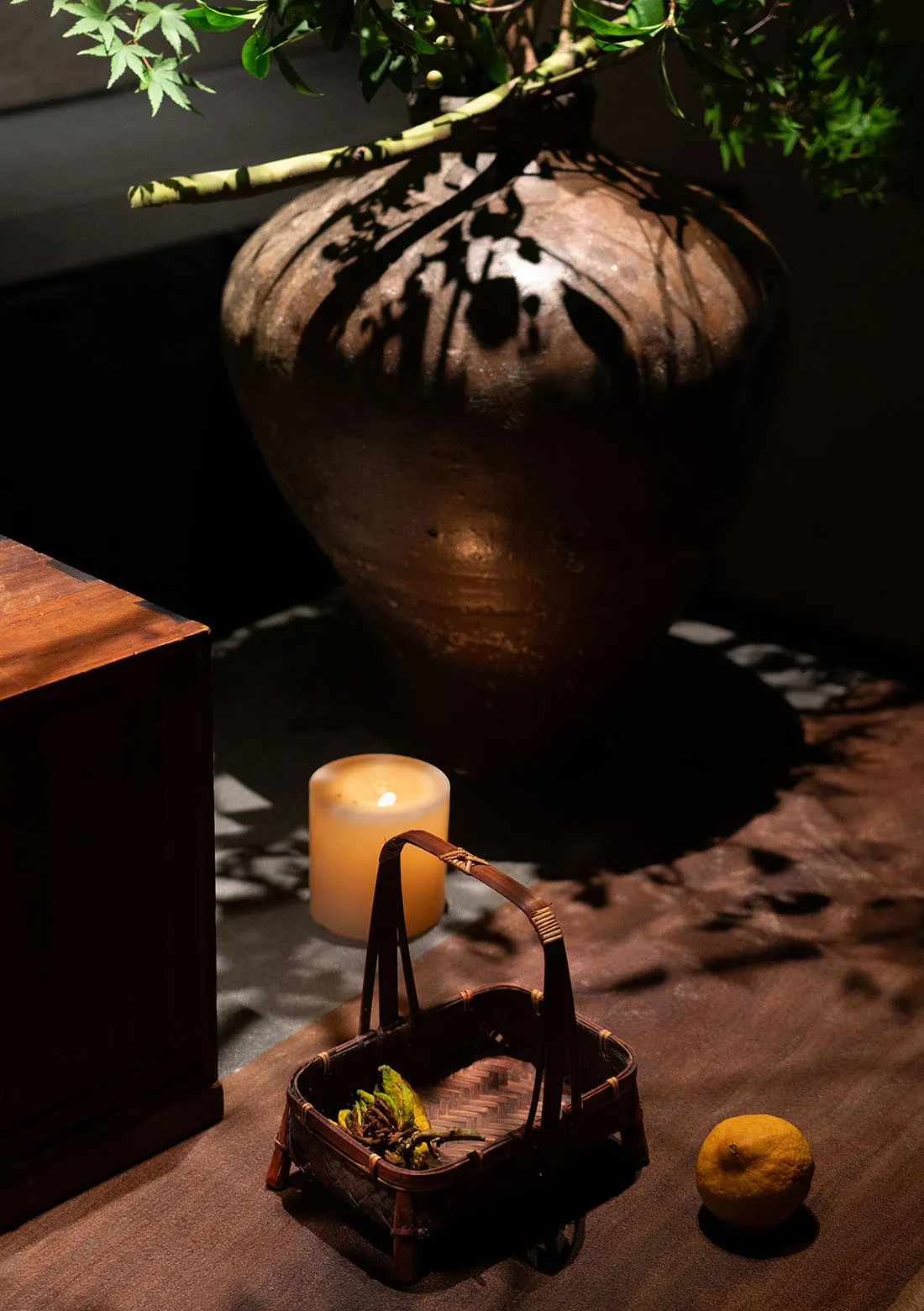 A ceramic vase casting a shadow on a wooden table with a lit candle and basket of leaves.