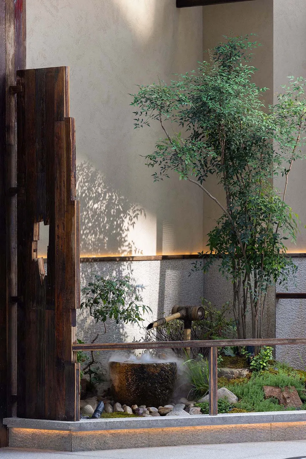 A modern indoor Japanese garden featuring a wooden wall, bamboo structure, stone basin, and lush green plants.