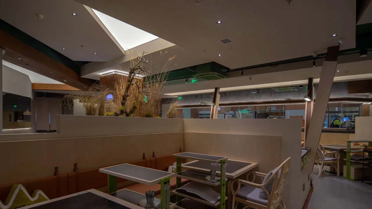 Modern restaurant interior with wooden tables, glass walls, and metal accents. Features an artificial plant centerpiece.